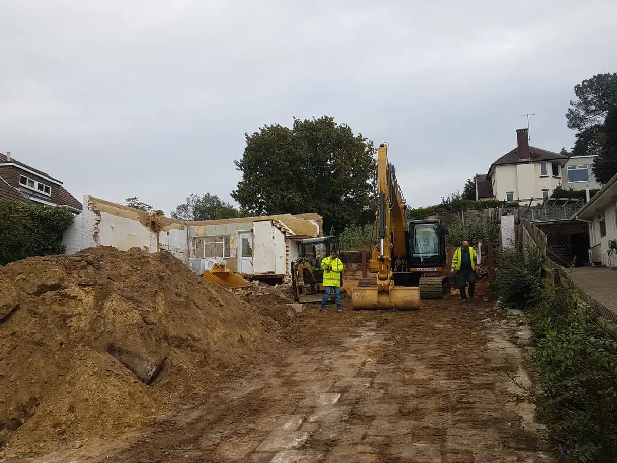 Partially demolished house with excavator, workers and muddy excavation